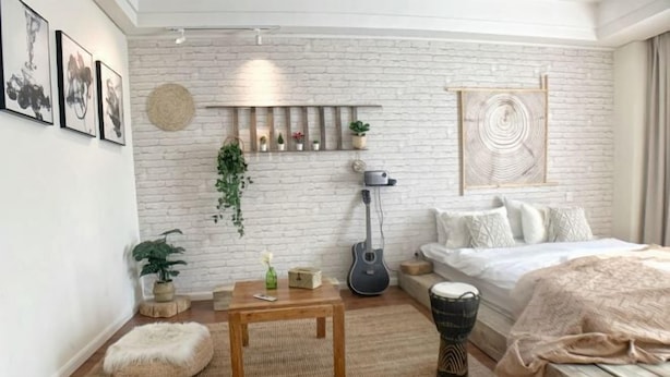Een moderne slaapkamer met een witte bakstenen muur, een bed met een wit en beige dekbedovertrek, een gitaar, een trommel en een klein houten tafeltje met een plant.