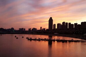 Een stadshemelbeeld bij zonsondergang, met gebouwen die silhouet vormen tegen de lucht, een kalme rivier met bootjes, en een heldere horizon.