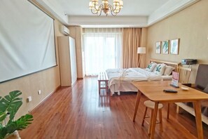 Habitación de hotel con piso de madera, una cama con ropa blanca, un escritorio con silla, una lámpara de pie y una pantalla de proyector.