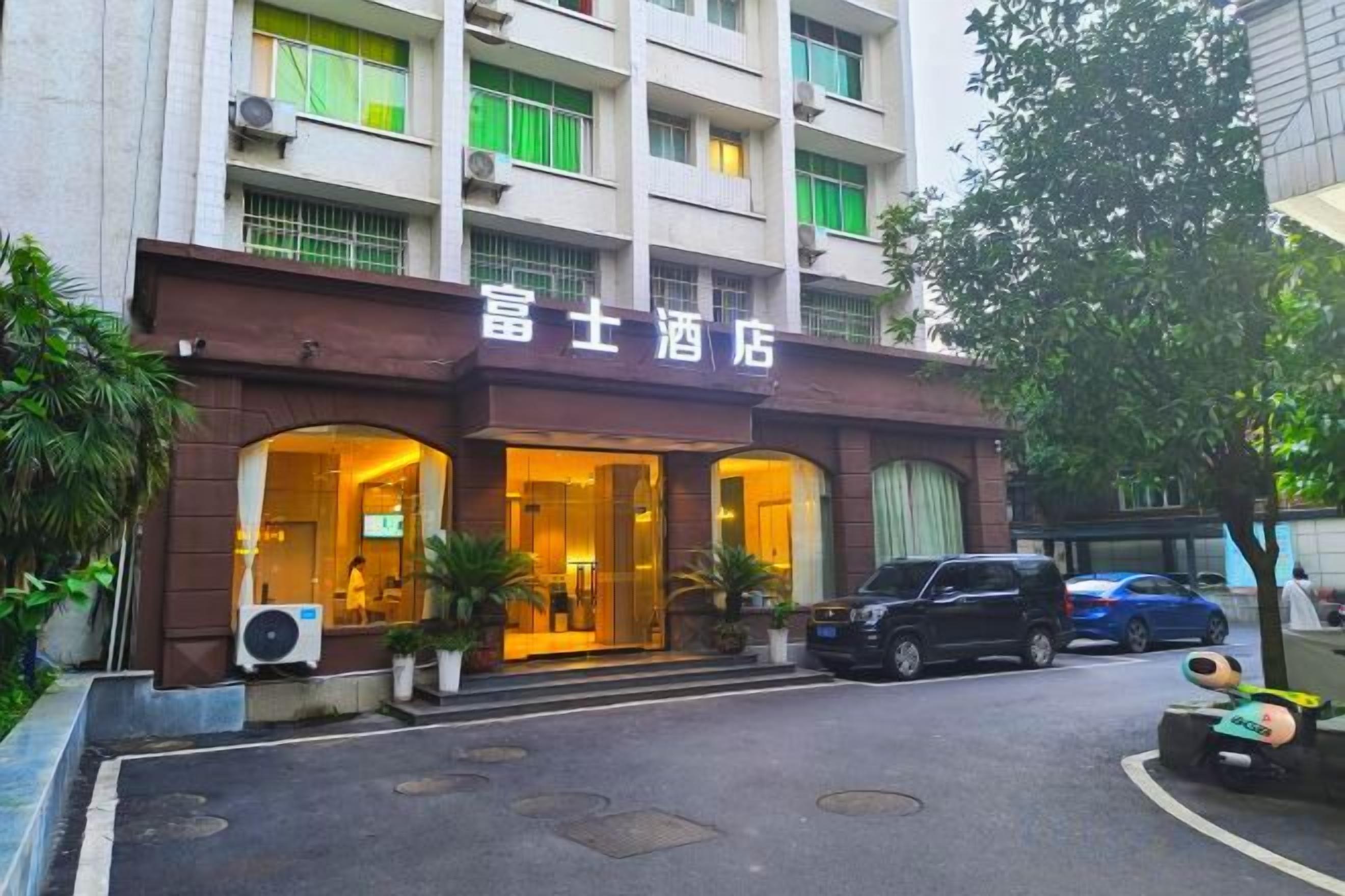 A hotel entrance with a brown facade, large windows, and a parking area in front.