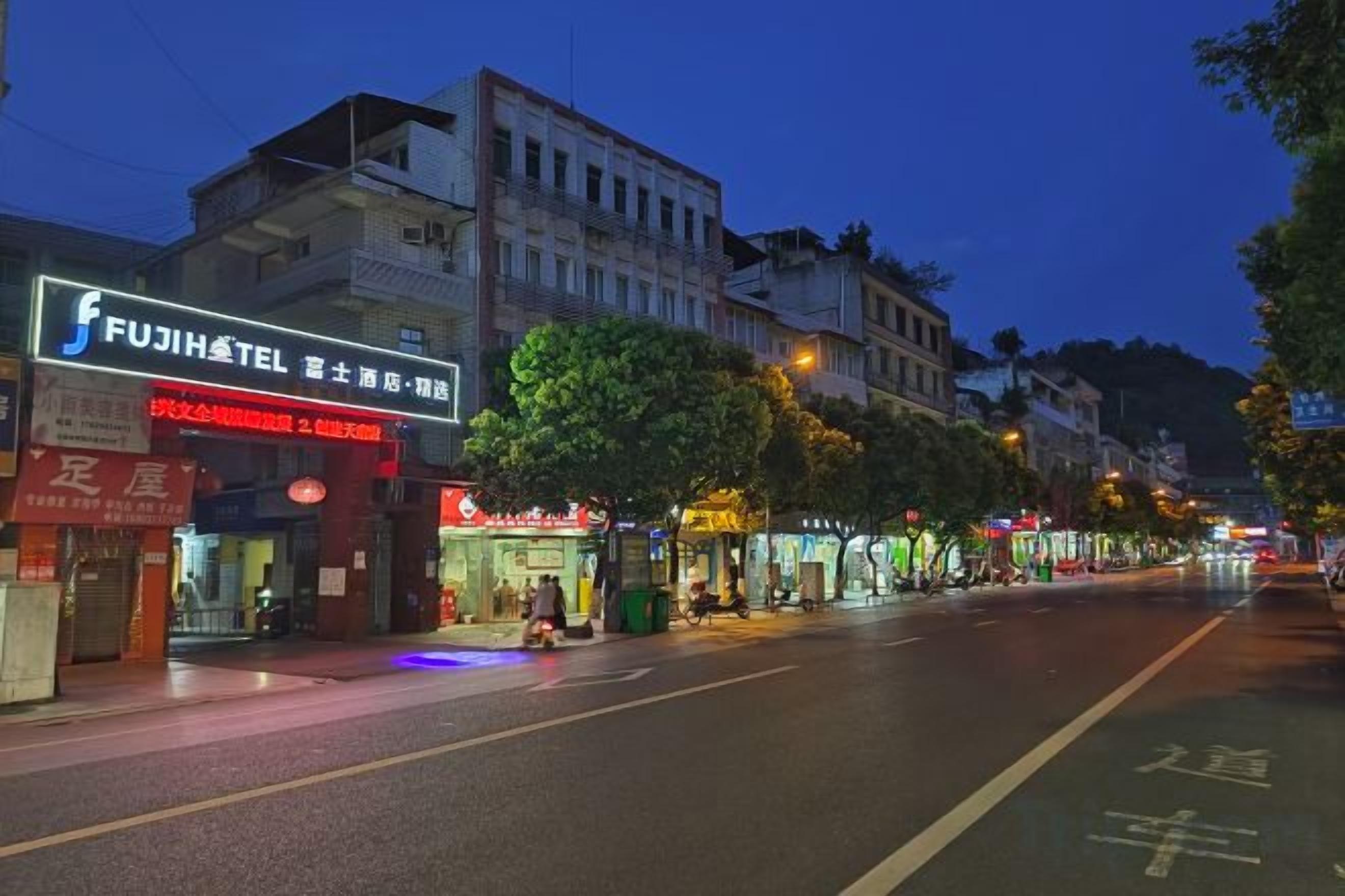 Un hotel llamado FUJIHOTEL con un letrero visible, una calle con tiendas y árboles bordeando la acera.