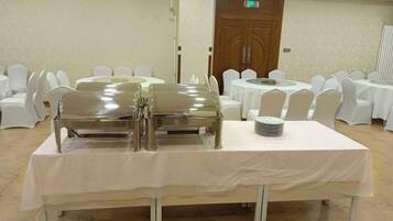 A banquet table with a white tablecloth, surrounded by white chairs, in a room with a wooden door and white walls.