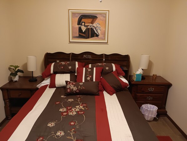 Una cama bien hecha con una colcha estampada con motivos florales, dos mesitas de noche con lámparas, una obra enmarcada sobre la cama y una pequeña planta en maceta a la izquierda.