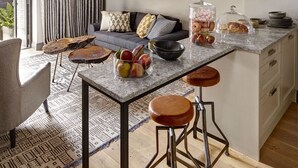 A modern kitchen with a central island, bar stools, a hanging light fixture, and a dining area with a sofa and curtains.