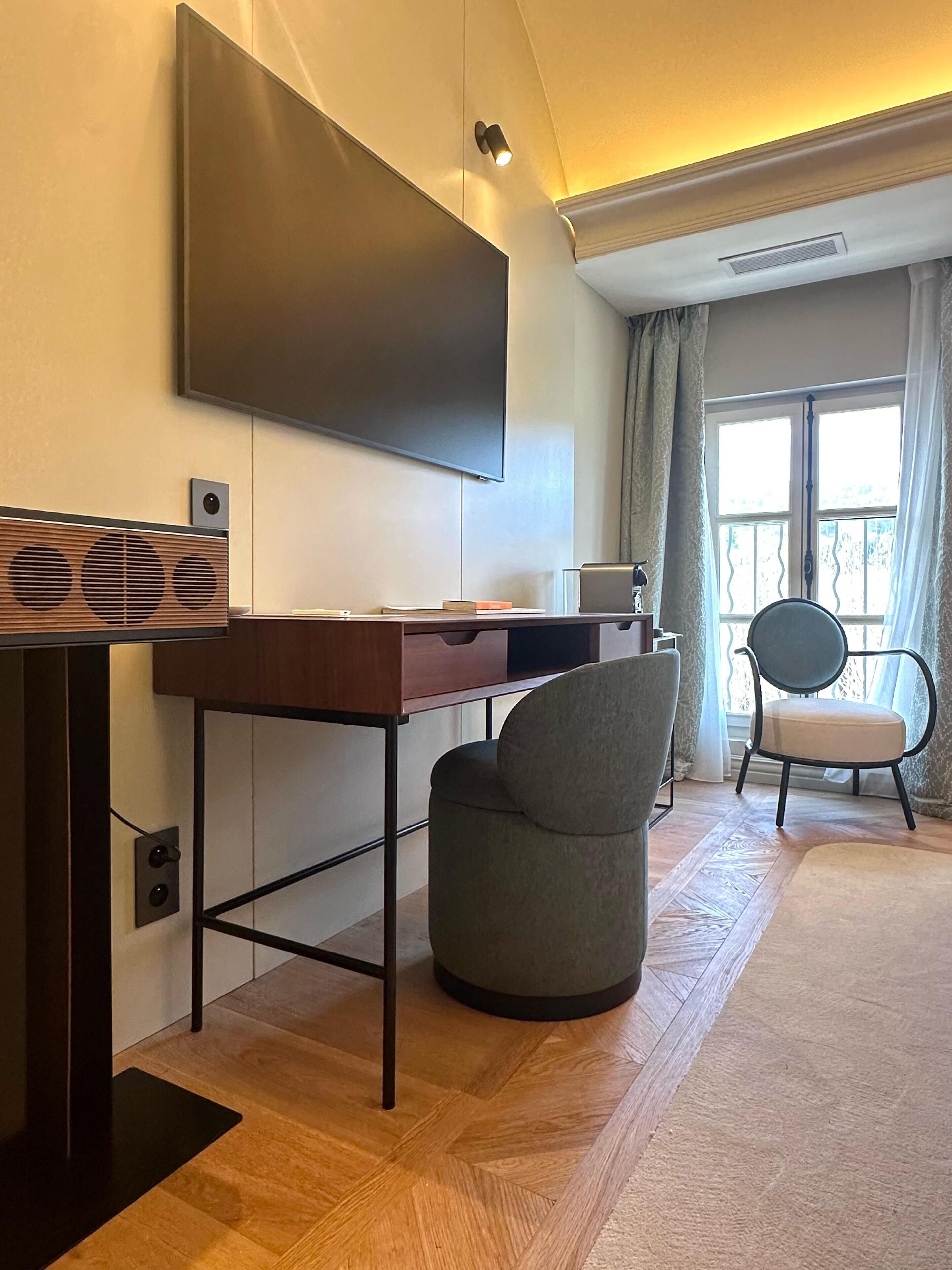 Una sala moderna con un televisor de pantalla plana montado en la pared, un escritorio de madera con una computadora, un sillón gris y una ventana con cortinas.