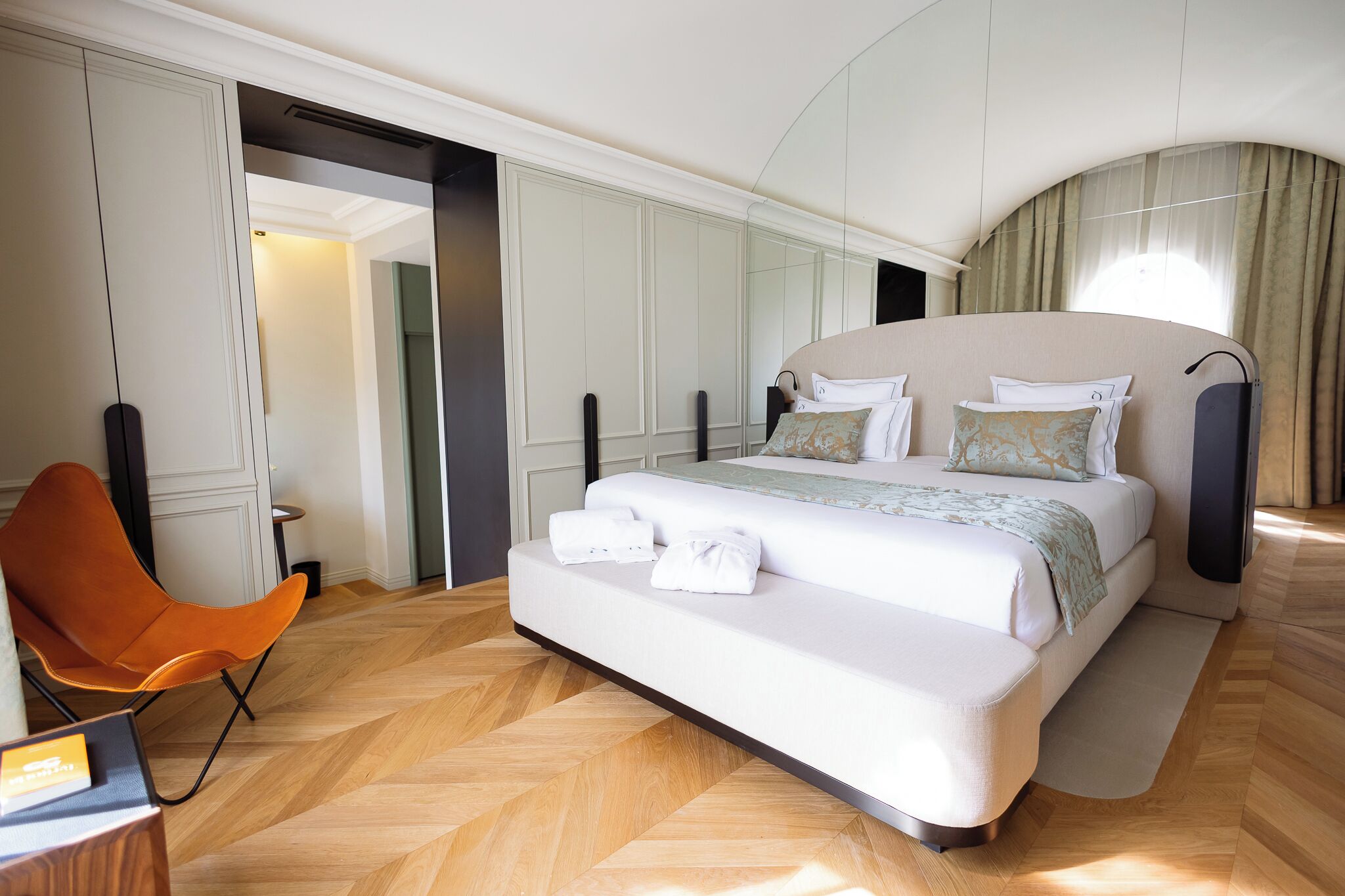 A modern hotel room with a large bed, a seating area with a chair, and a mirror wall.