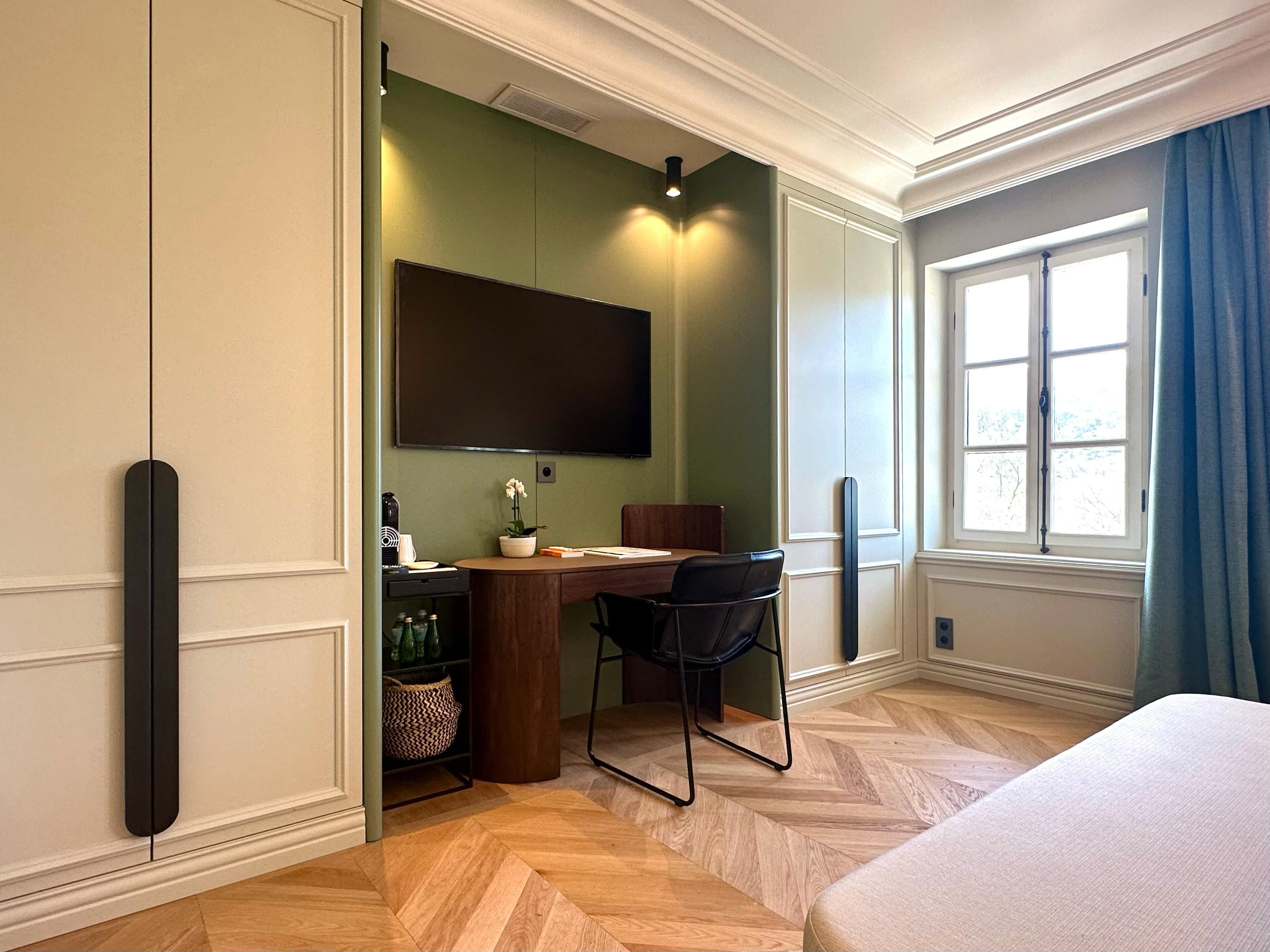 A hotel room with a wooden herringbone floor, a desk with a chair, a bed, a large window with curtains, and a mounted flat-screen TV.