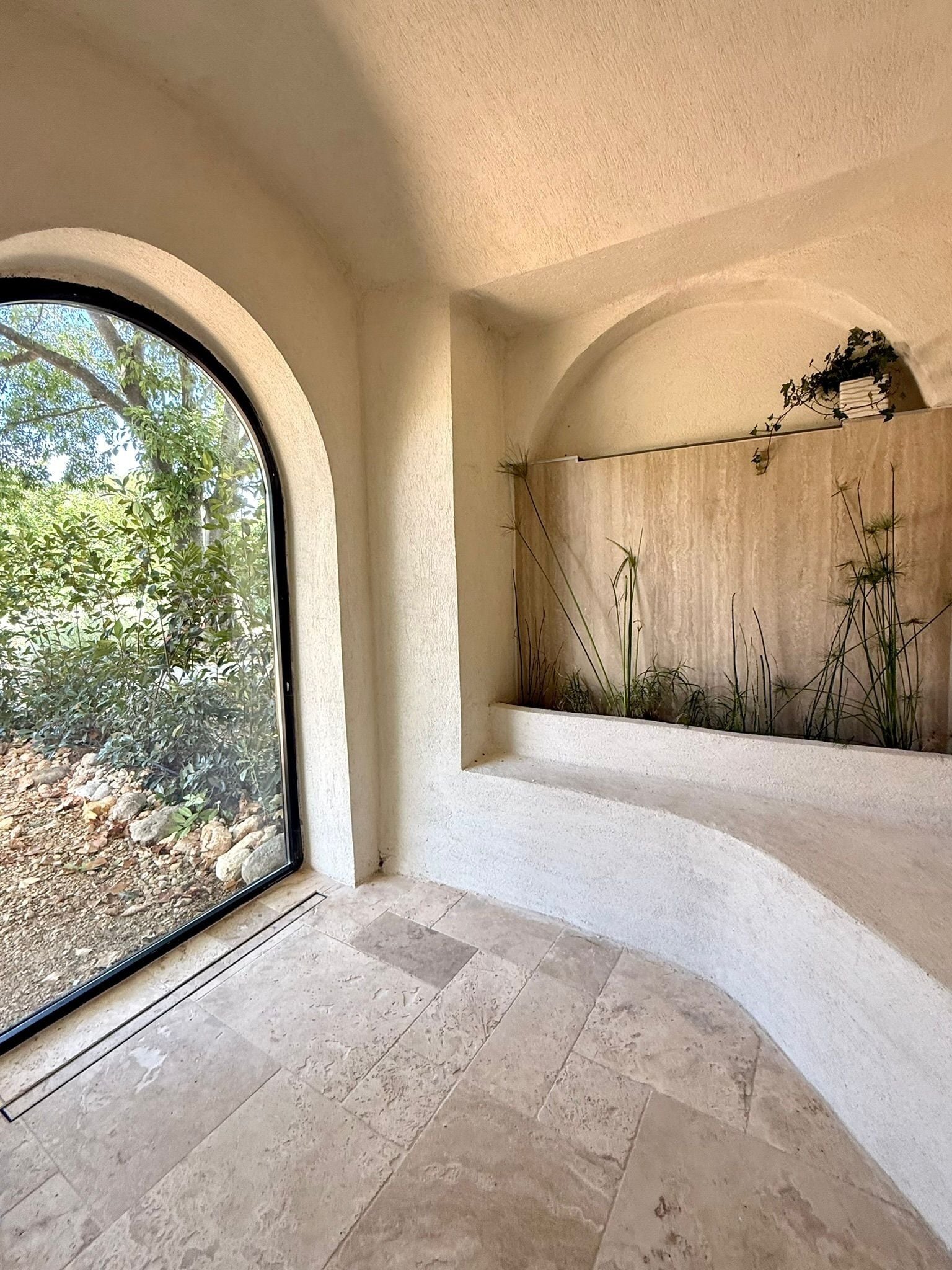 Une pièce dotée d’une grande fenêtre en arc de cercle, d’un jardin intégré où poussent de hautes plantes et d’un sol recouvert de carreaux de pierre.