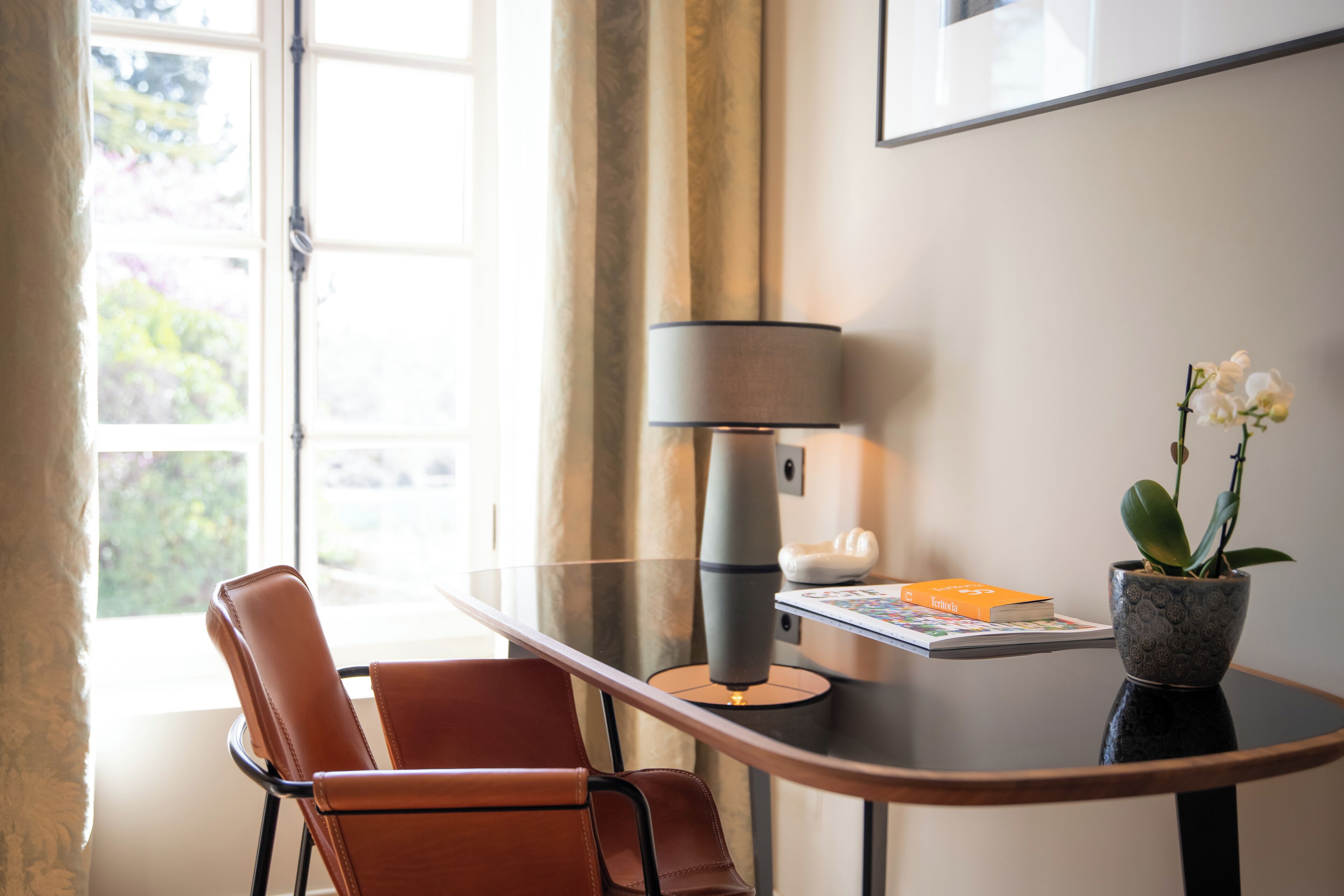 Una habitación moderna con una mesa de vidrio, un sillón de cuero marrón, una lámpara, una orquídea en maceta y un cuadro en la pared.