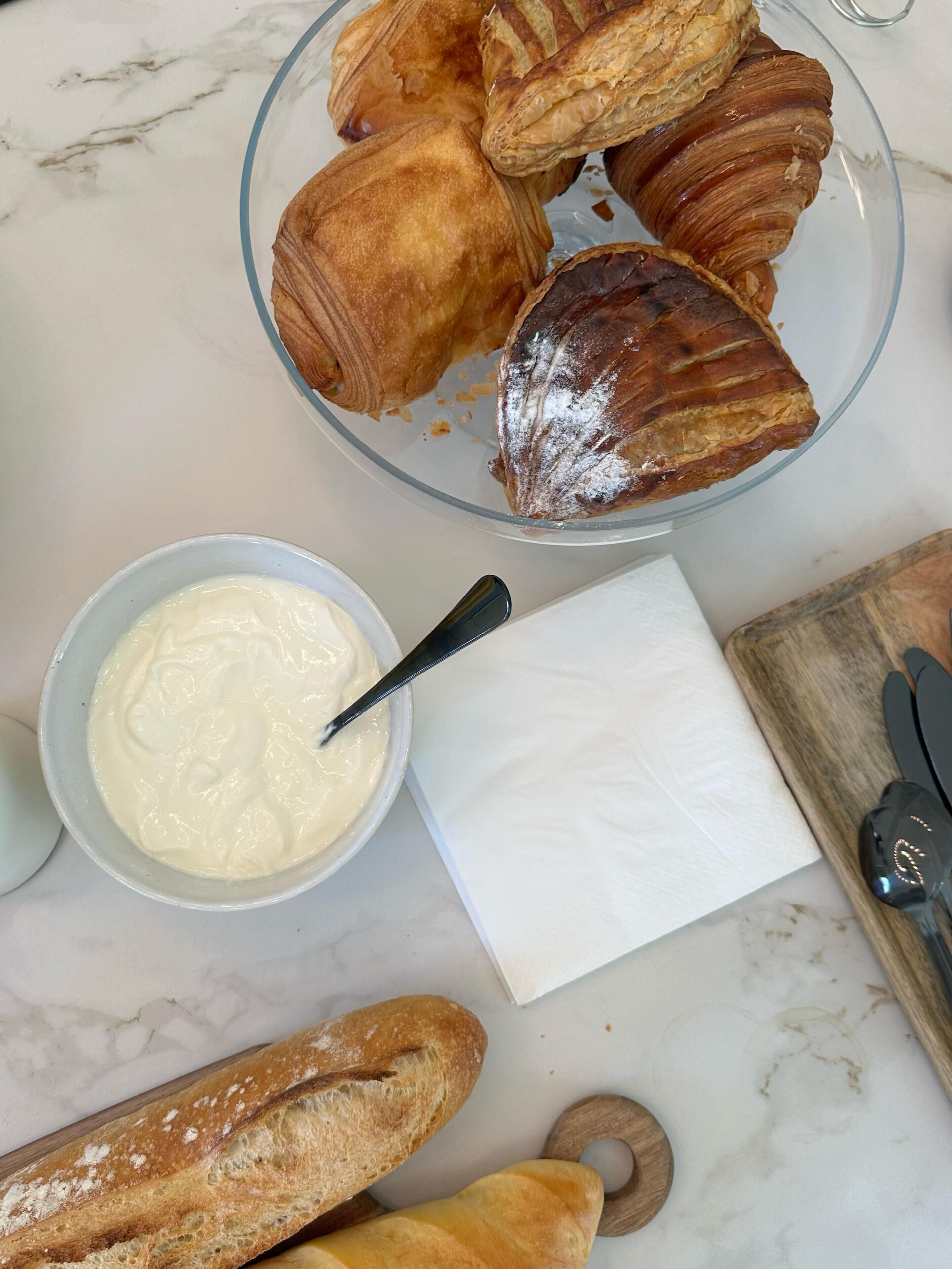 A marble countertop with a glass bowl of croissants, a bowl of cream, a napkin, a loaf of bread, and a wooden cutting board with a knife.