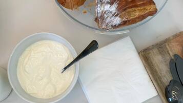 A marble countertop with a glass bowl of croissants, a bowl of cream, a napkin, a loaf of bread, and a wooden cutting board with a knife.