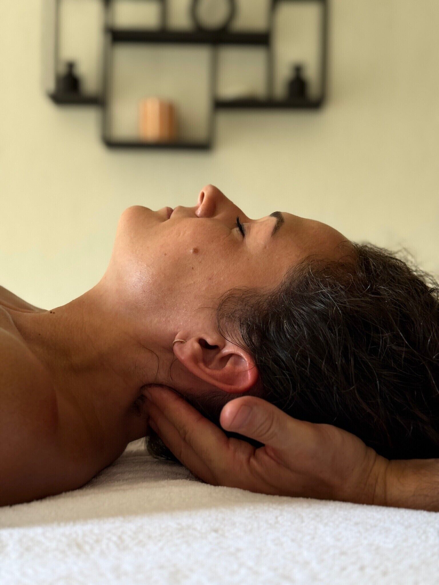 A person lying on a white towel with a hand resting on their head, in a room with a plain wall and a shelf with items.