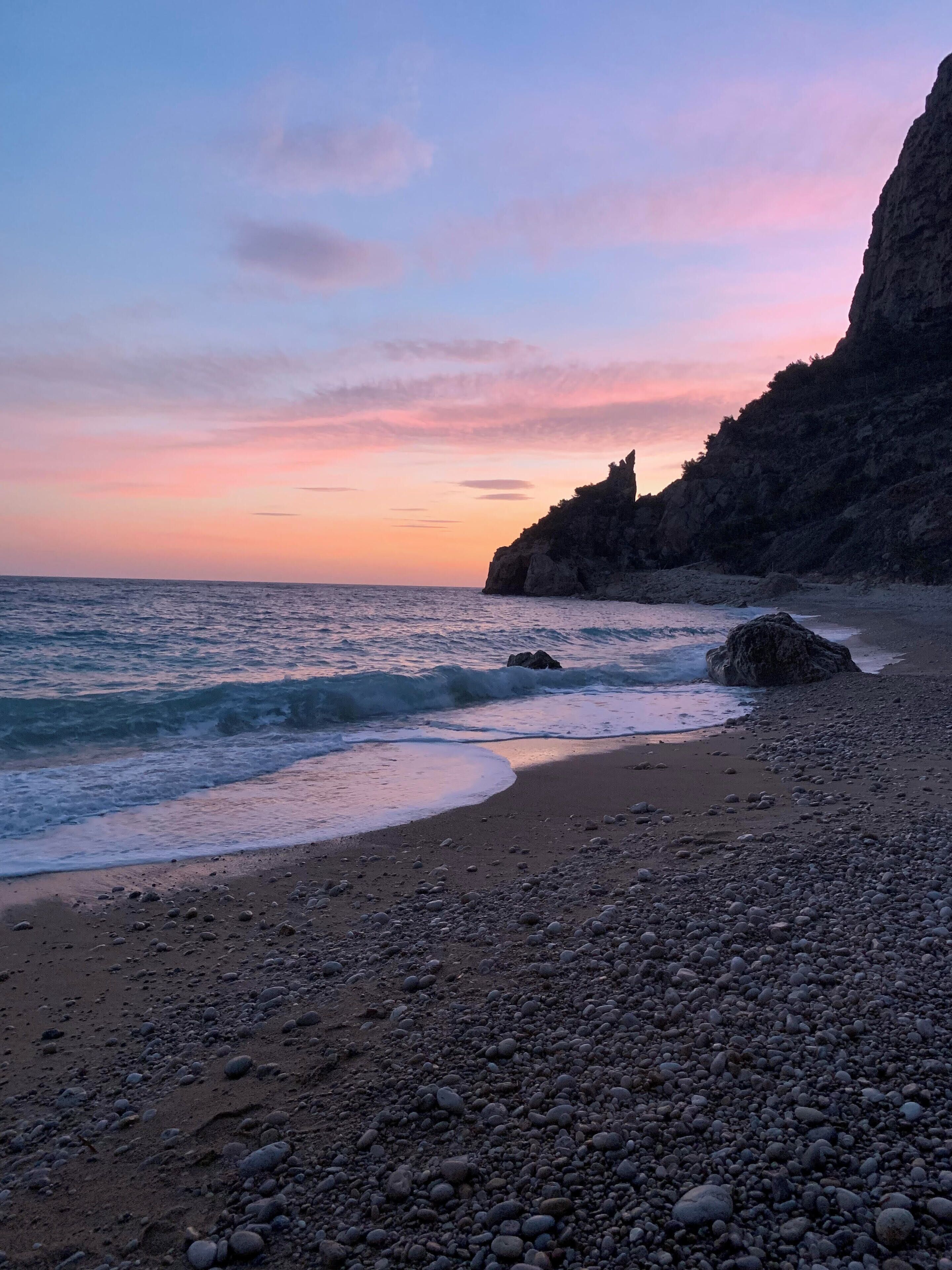 Ein Kiesstrand mit Wellen im Abendrot, felsige Klippen und ein klarer Himmel.