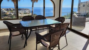 A dining area with a glass table and wicker chairs, overlooking a coastal view with buildings and palm trees. - Magnificent Villa with swimming pool and panoramic view, ideal for 6 people. (Cumbre del Sol)