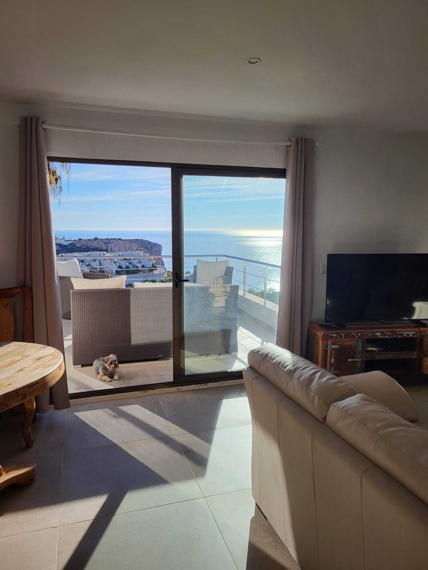 Una sala de estar con sofá, televisor y balcón con vista al mar y edificios.