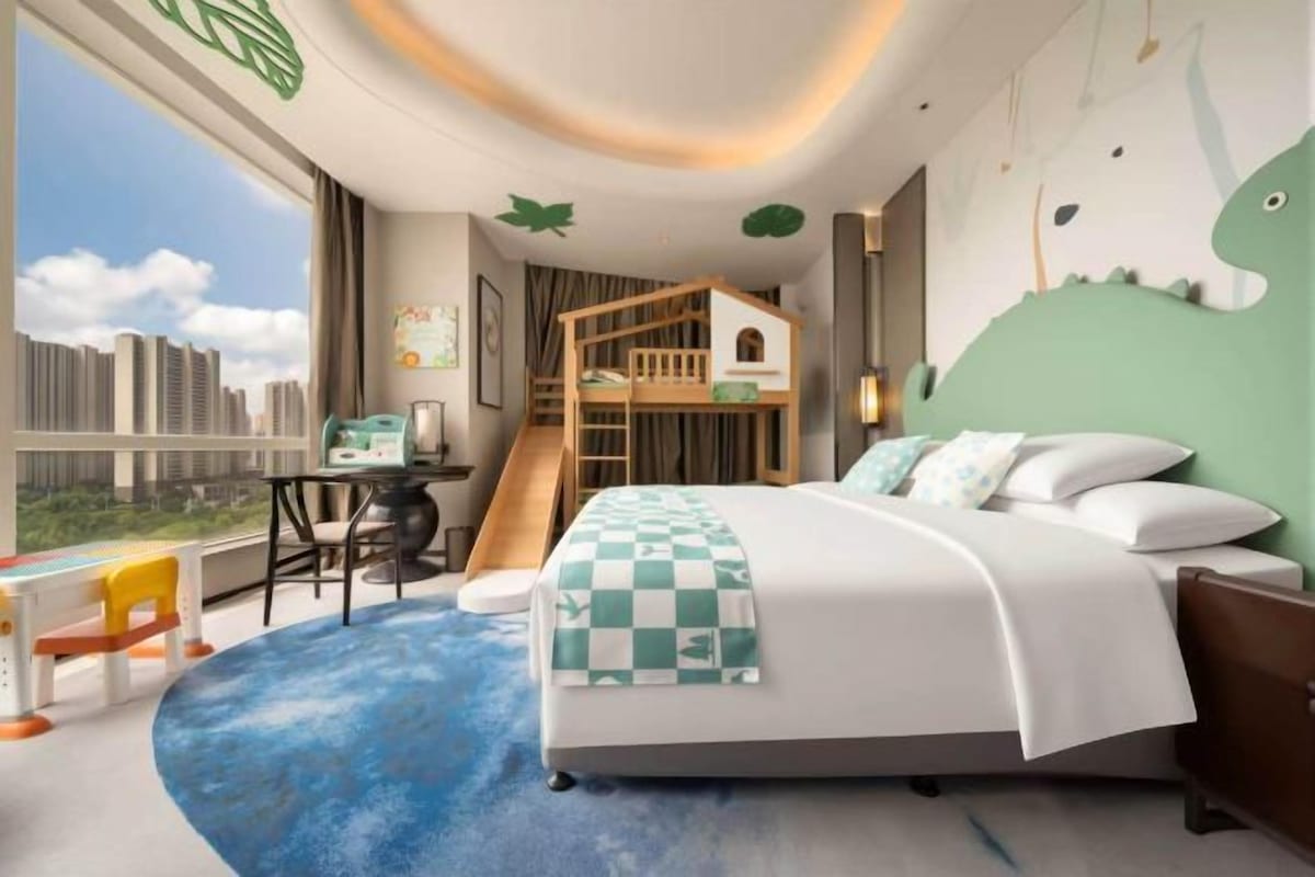 A hotel room with a bunk bed, a desk with a computer, a view of the city, and a large window.