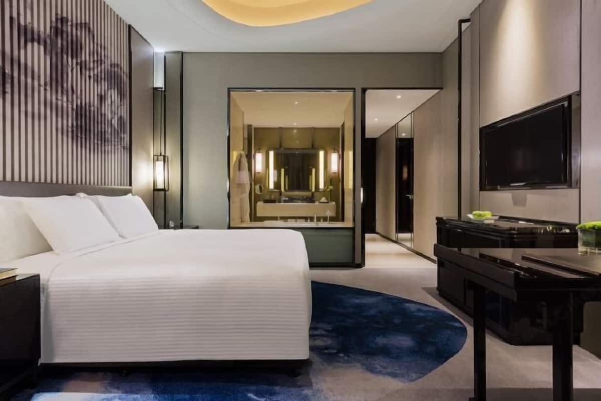 A modern hotel room with a large bed, a desk, a flat-screen TV, and a bathroom with a mirror and lights.