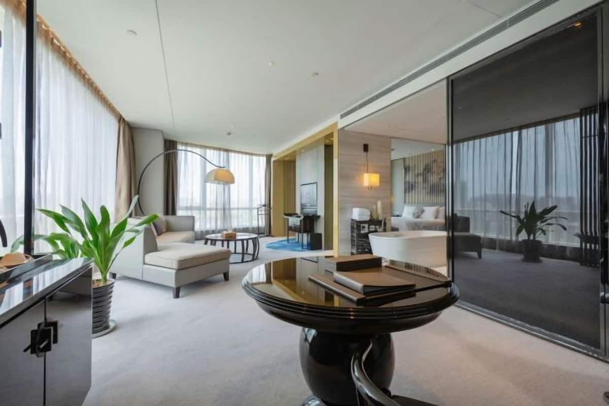 A modern hotel room with a large window, a round black table, a sofa, and a desk area.