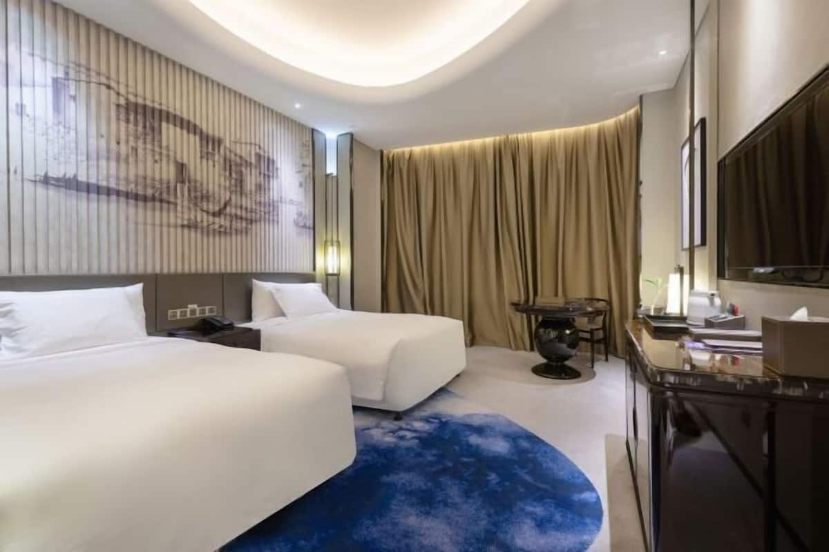 A hotel room with two beds, a desk, a chair, a TV, and a large artwork on the wall.