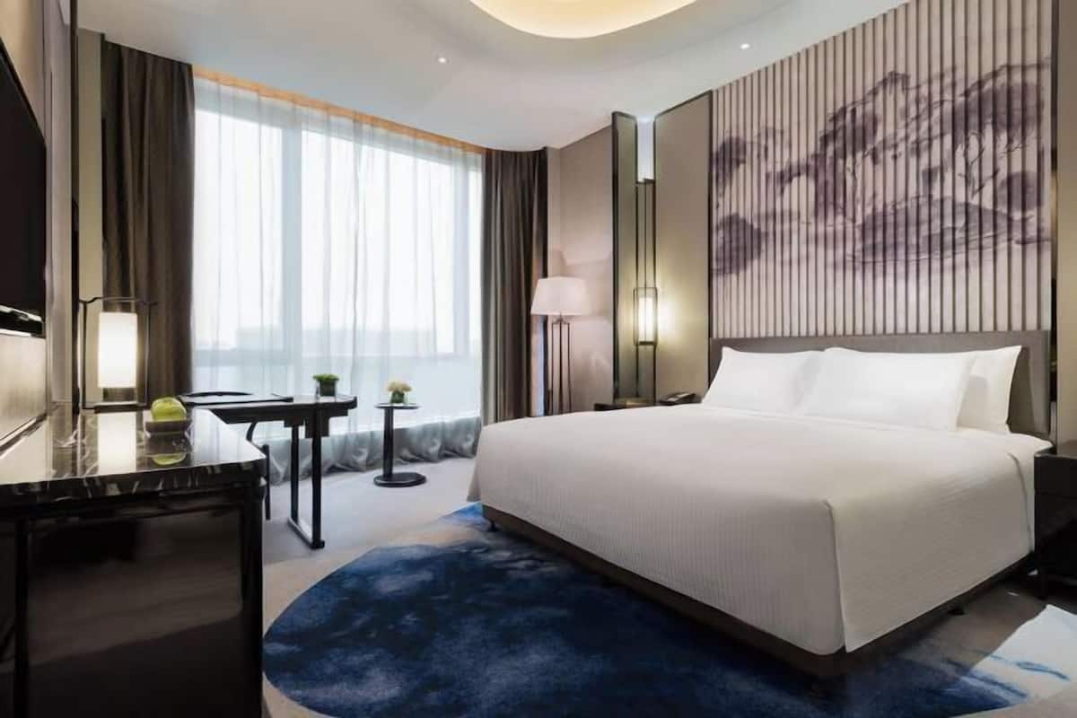 A hotel room with a large bed, a desk, a chair, a lamp, a small table with a plant, and a large window with curtains.