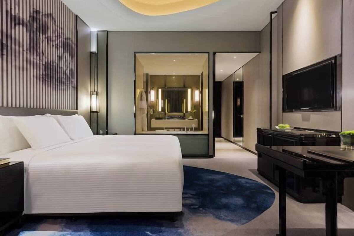 A modern hotel room with a large bed, a desk, a TV, and a bathroom with a mirror and lights.