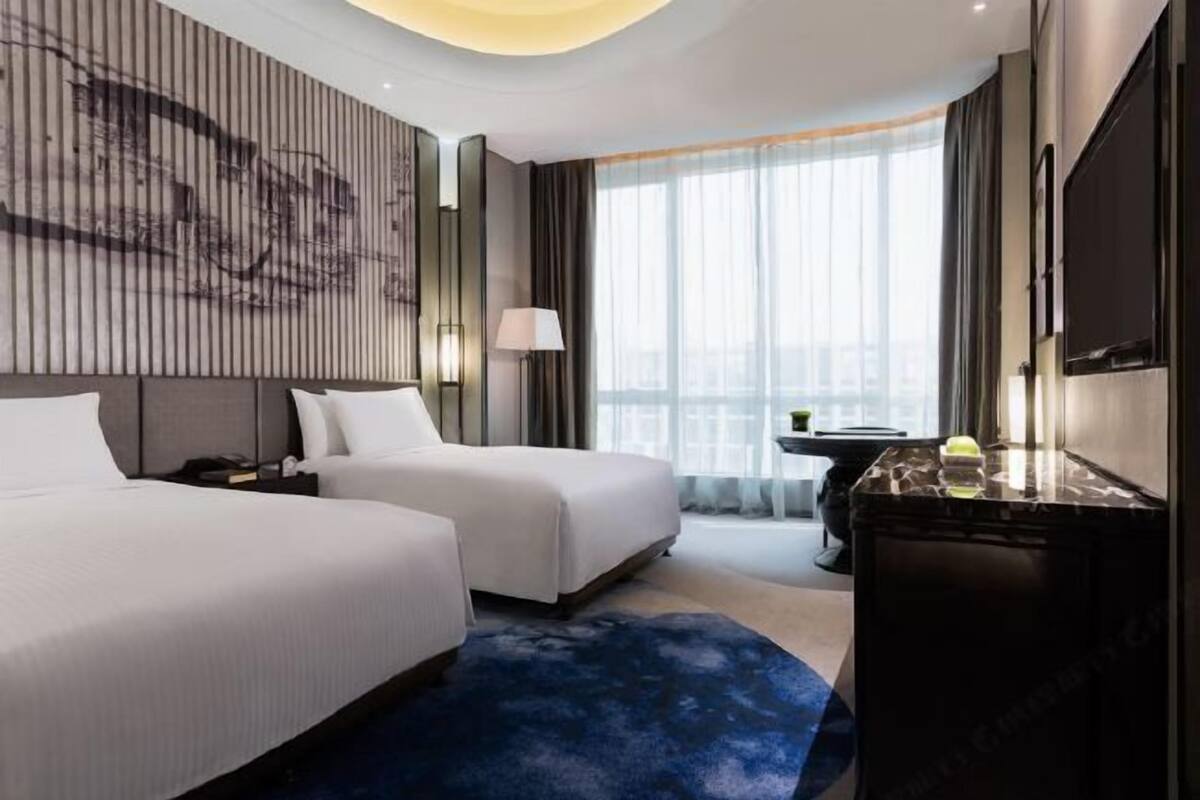 A hotel room with two beds, a desk, a lamp, a TV, and a large window with curtains.