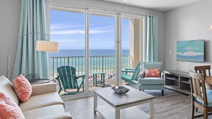 Un vaste salon doté d’une grande porte-fenêtre coulissante donnant sur un balcon avec vue sur l’océan. On y trouve deux fauteuils verts, un canapé blanc et une petite table sur laquelle est posé un vase décoratif.
