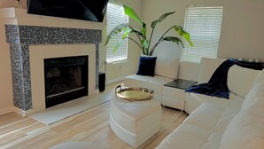 A modern living room with a fireplace, white sofas, a flat-screen TV, and large windows with blinds. - Backyard Oasis! (San Antonio)