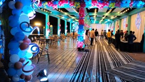 Un espacio interior amplio con filas de mesas y sillas, decorado con globos de colores y luces colgantes.