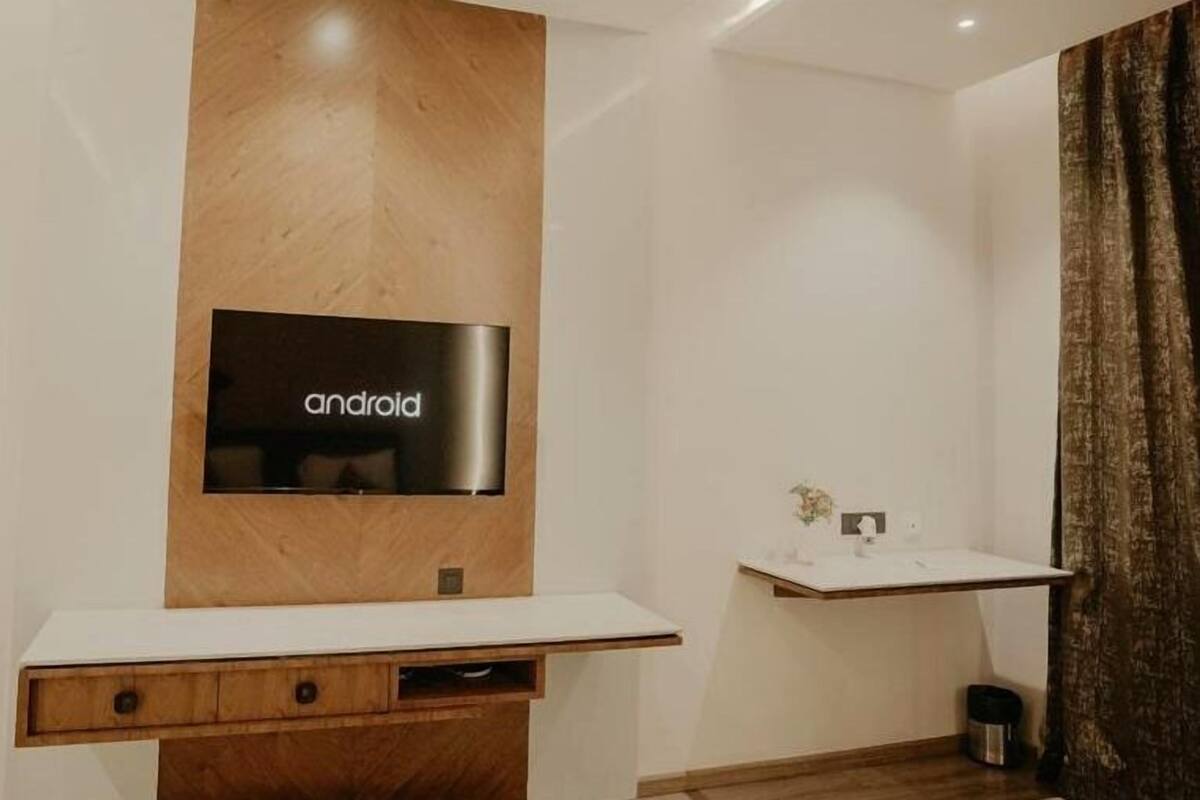 A modern bathroom with a wooden vanity, a mounted television displaying 'android', and a small shelf with a vase.