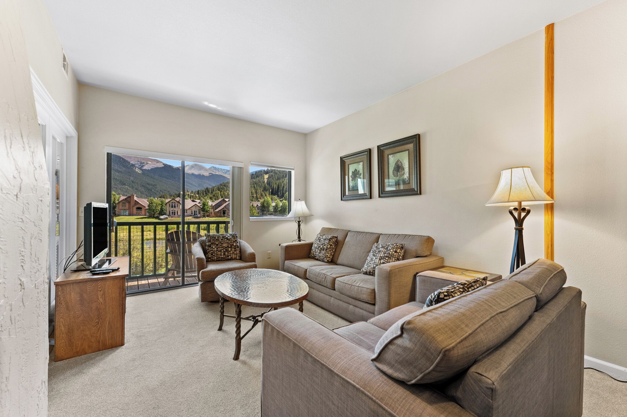 A living room with a sofa, a coffee table, a TV, and a lamp. There are two windows with a view of a mountain and houses.