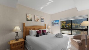 A hotel room with a large bed, a wooden headboard, a nightstand with a lamp, a chair, and a window with a view of mountains and a lake. - Gac217c1 (Copper Mountain)