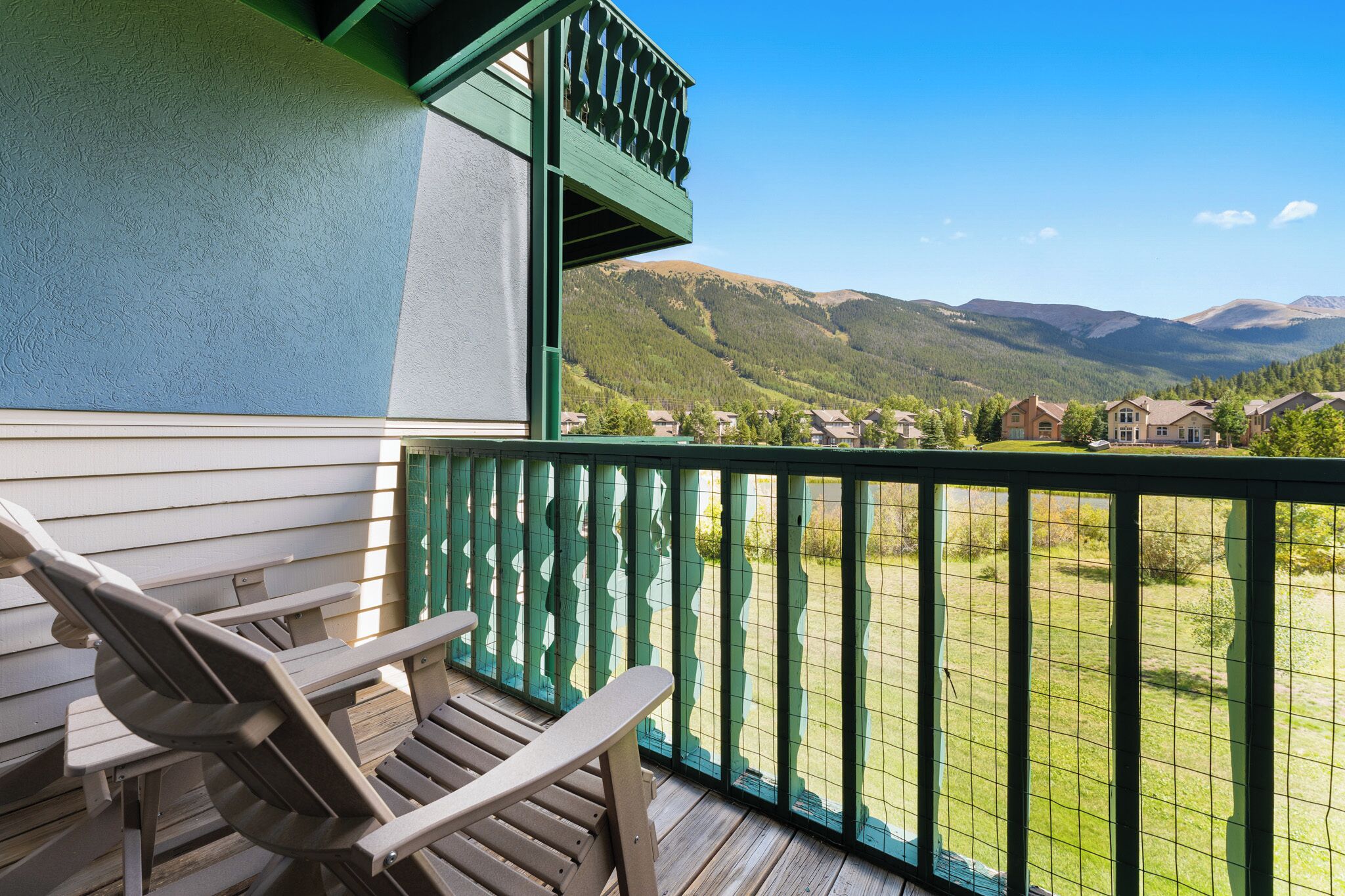 A wooden deck with a green railing, overlooking a mountainous landscape with houses in the distance.