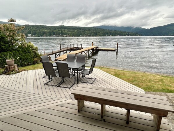 A wooden deck with a table and chairs overlooking a lake, a dock extending into the water, and a forested hillside in the background. - Quaint Lakefront Cottage w/Hot tub In Bellingham (Bellingham)