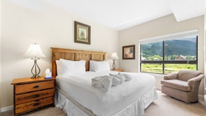 A bedroom with a large bed, two bedside tables, a chair, and a window with a view of greenery. - Gac219c1 (Copper Mountain)