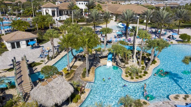An aerial view of a resort with a large swimming pool, palm trees, and a thatched-roof hut.