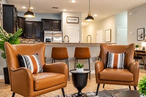 A modern kitchen with dark cabinets, a stainless steel refrigerator, and pendant lighting. There are two leather armchairs with patterned cushions, a small round table with a plant, and a rug on the floor.