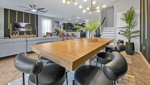 A modern living room with a wooden dining table, black leather chairs, a ceiling fan, and a large TV.