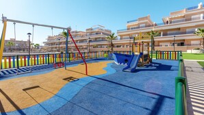 A playground with swings and slides, surrounded by a fence and palm trees, with apartment buildings in the background. - Beautiful home in Torre de la Horadada (Torre de la Horadada)