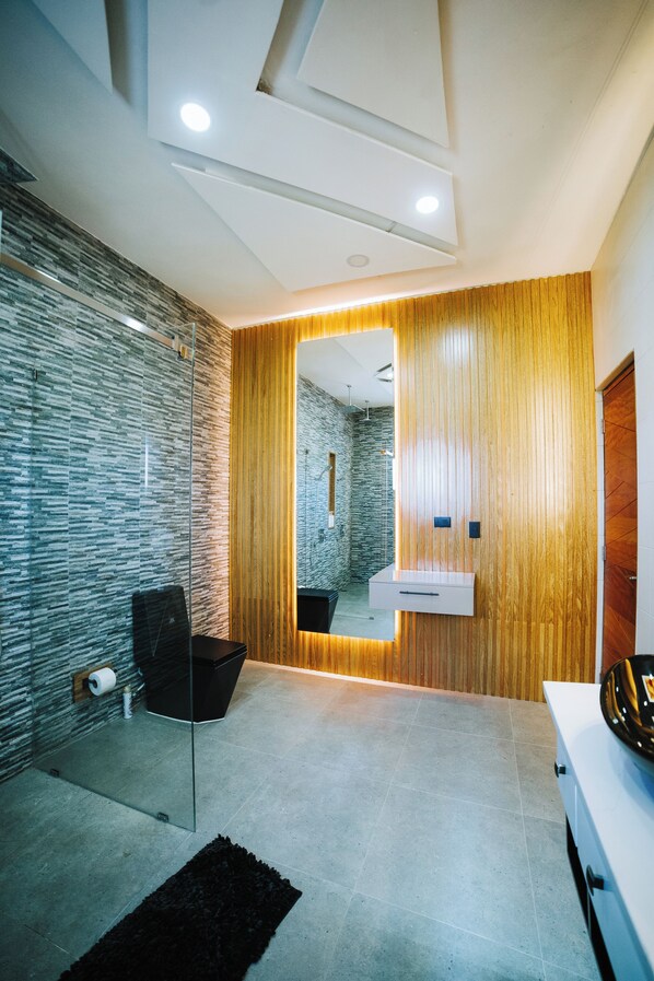 A modern bathroom with a glass partition, a wooden panel wall, a white sink, and a black trash bin.