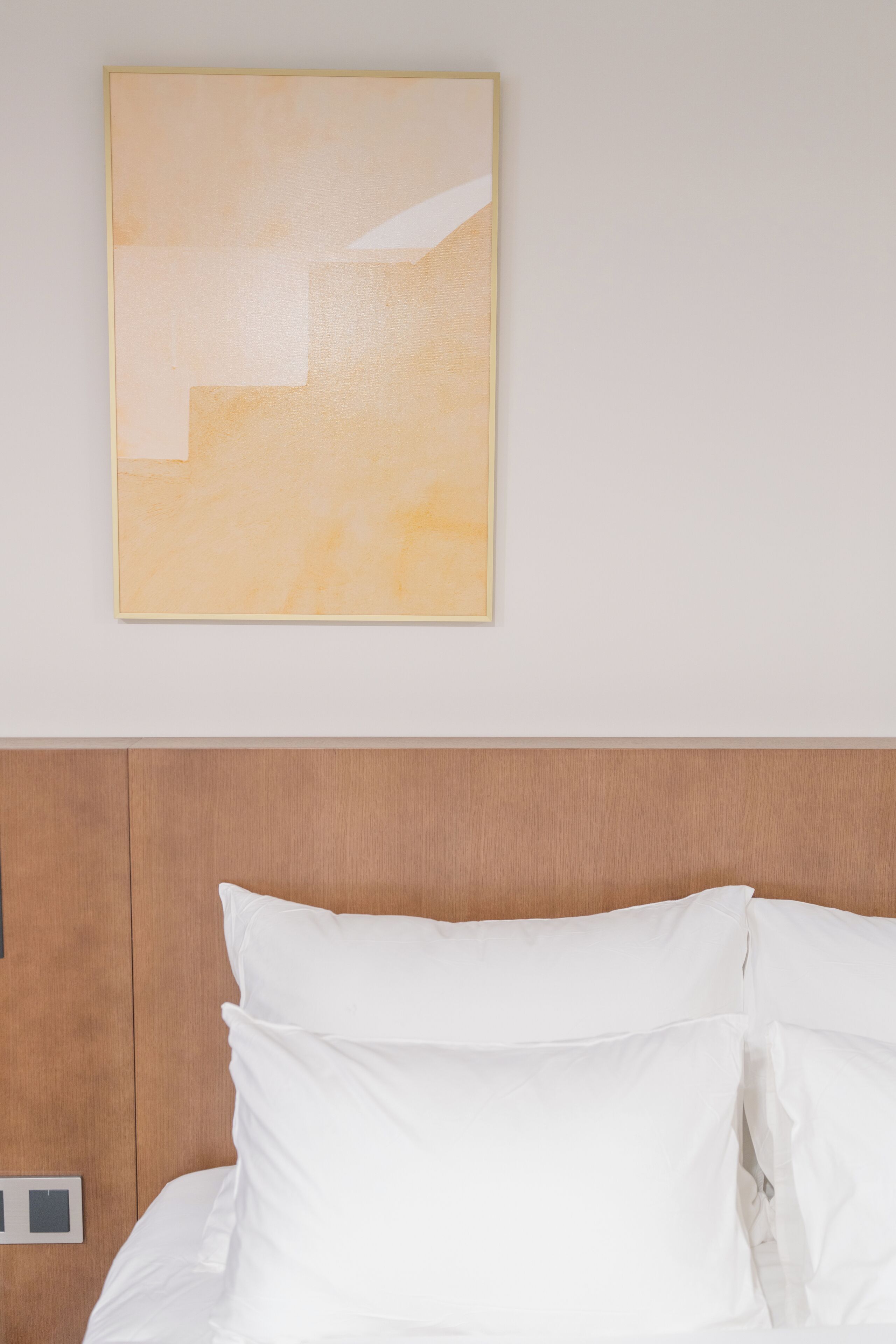 Una cama bien tendida con sábanas y almohadas blancas, un cabecero de madera y un cuadro abstracto colgado en la pared.
