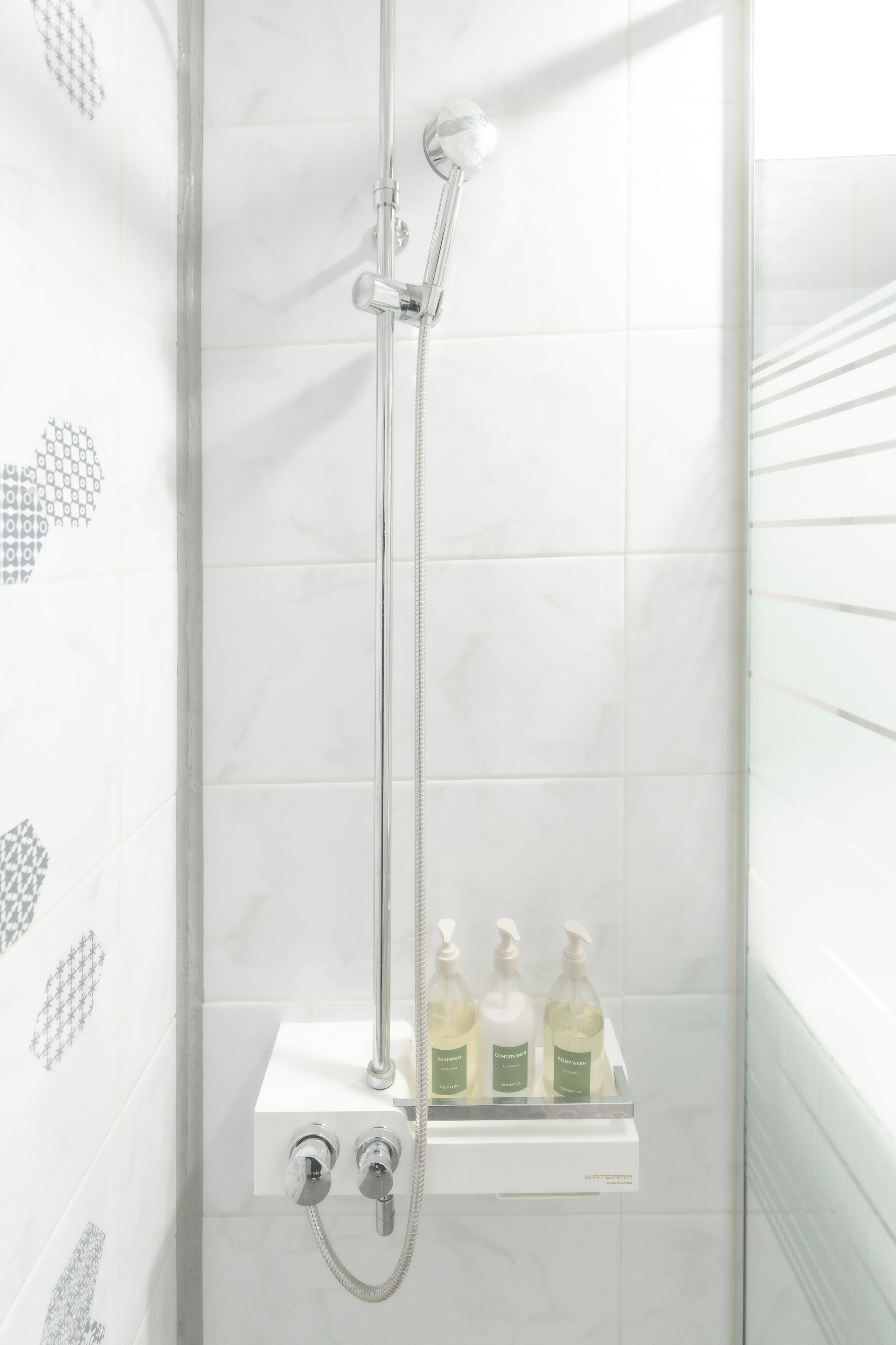 A shower with a chrome handle and a glass enclosure, a white tiled wall, and a shelf with three bottles of liquid soap.