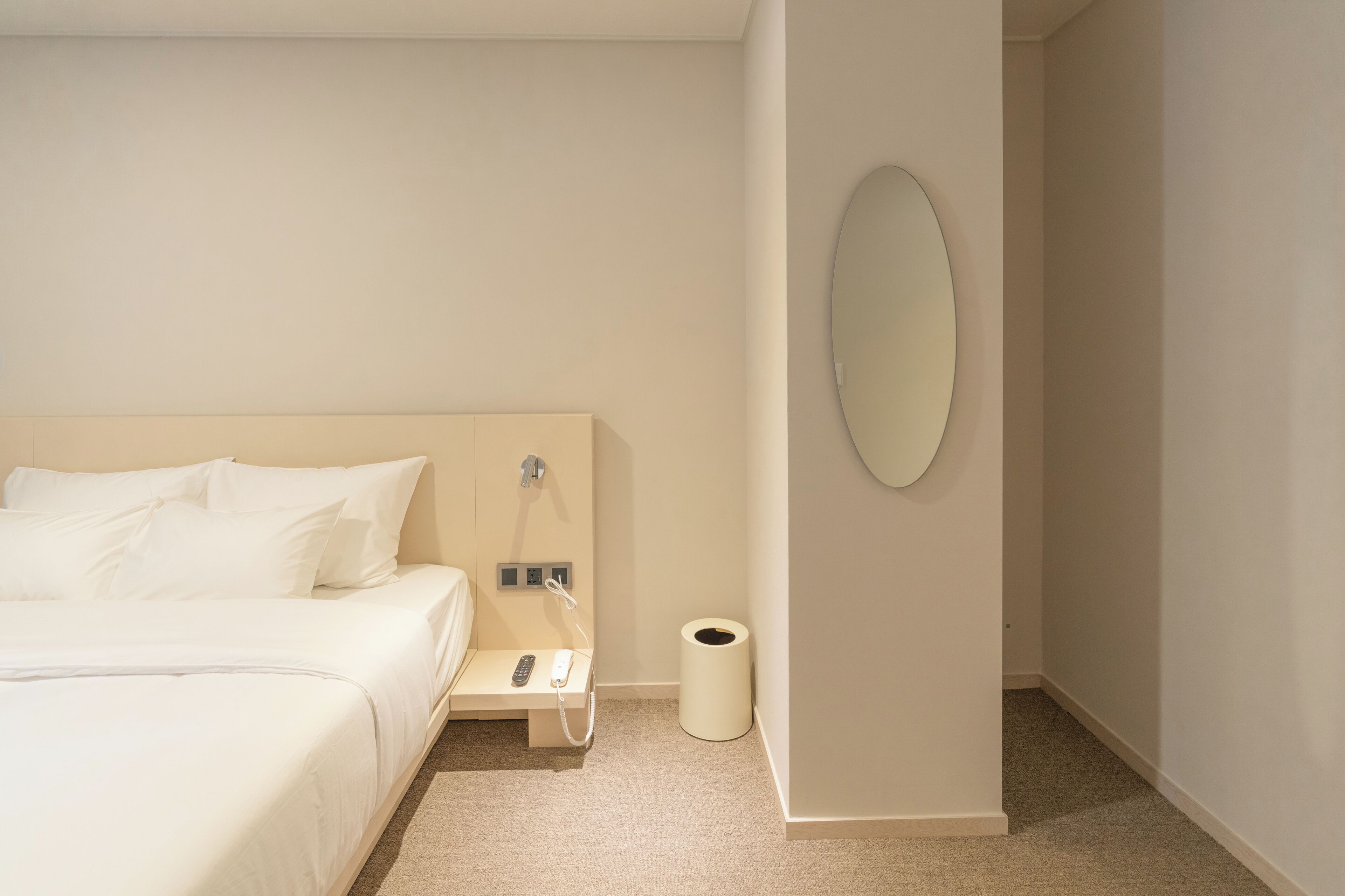 A hotel room with a bed, a bedside table with a phone, a trash bin, and a wall-mounted mirror.