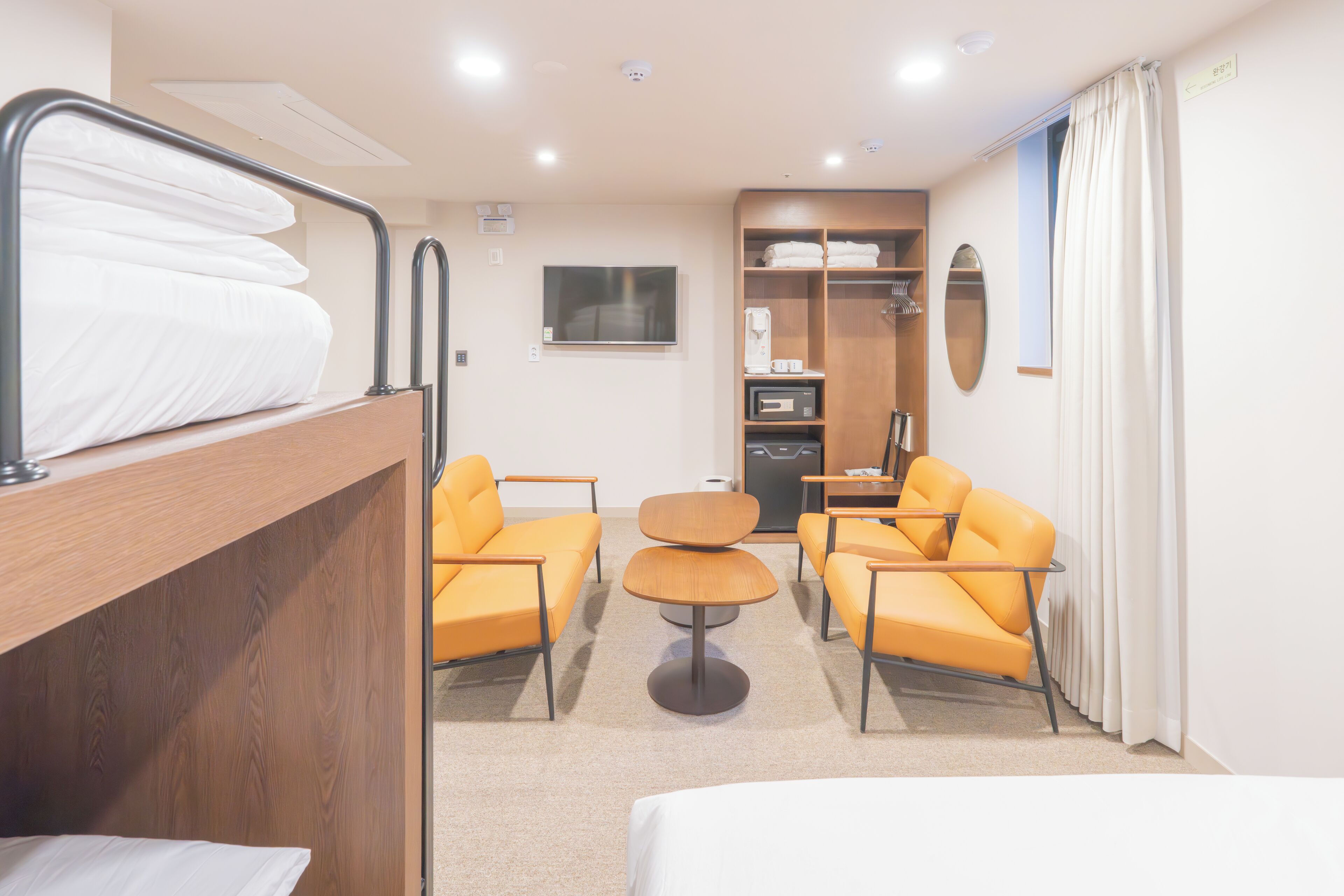 A hotel room with a bunk bed, two orange armchairs, a small round table, a TV, a wardrobe, and a window with curtains.