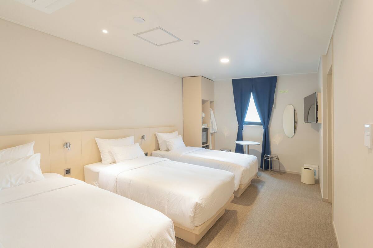 A hotel room with three single beds, a small table, a chair, a mirror, and a window with curtains.