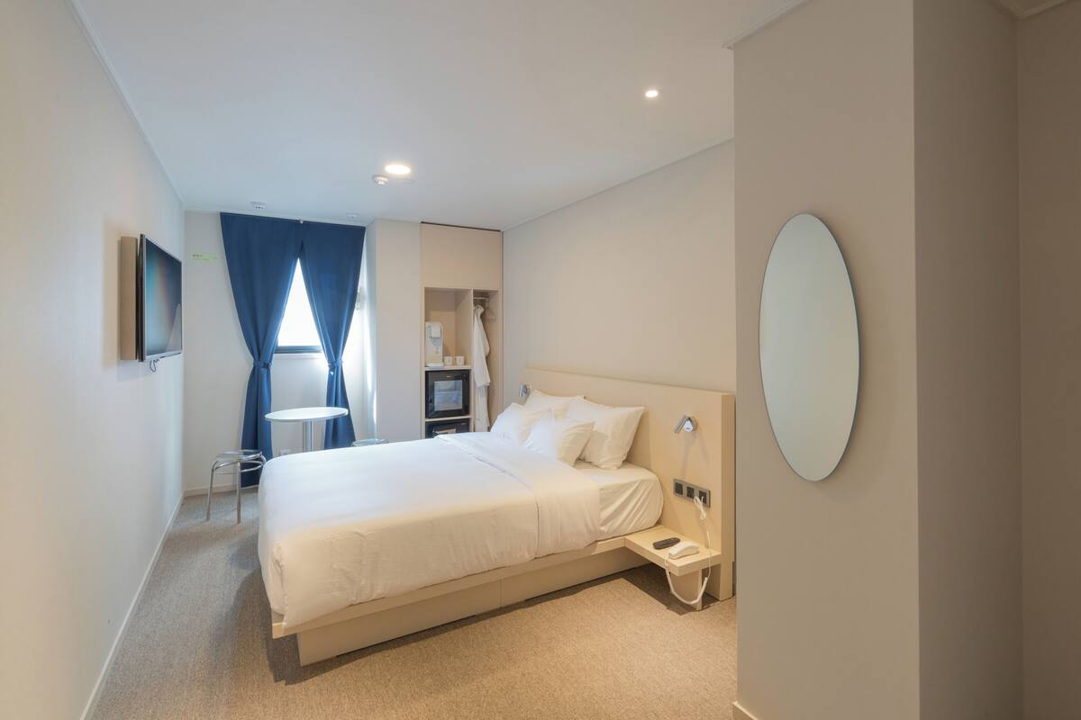 A modern hotel room with a large bed, a bedside table with a phone, a small desk with a chair, a TV, and a mirror on the wall.