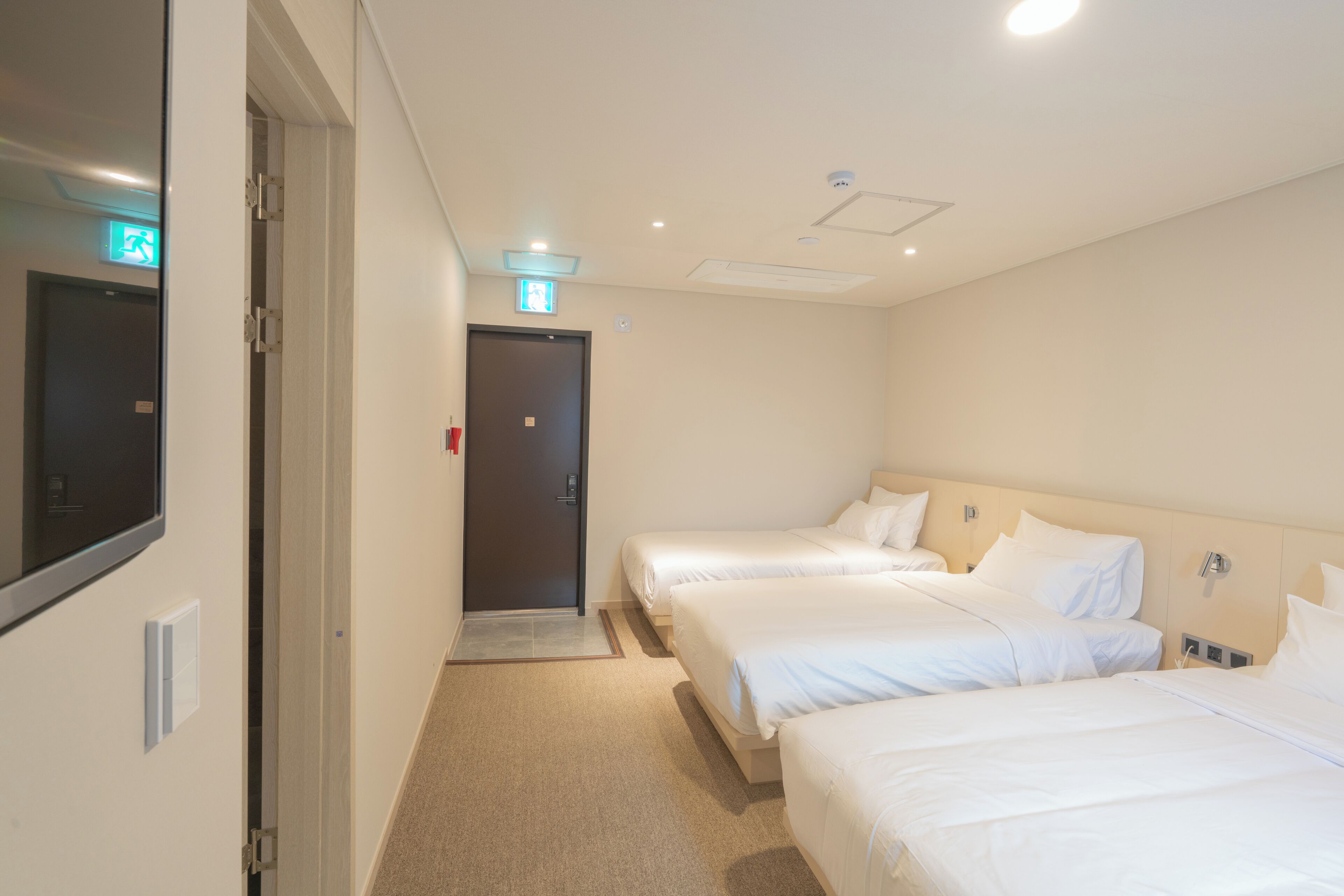 A hotel room with three single beds, a door with an emergency exit sign, and a light switch on the wall.