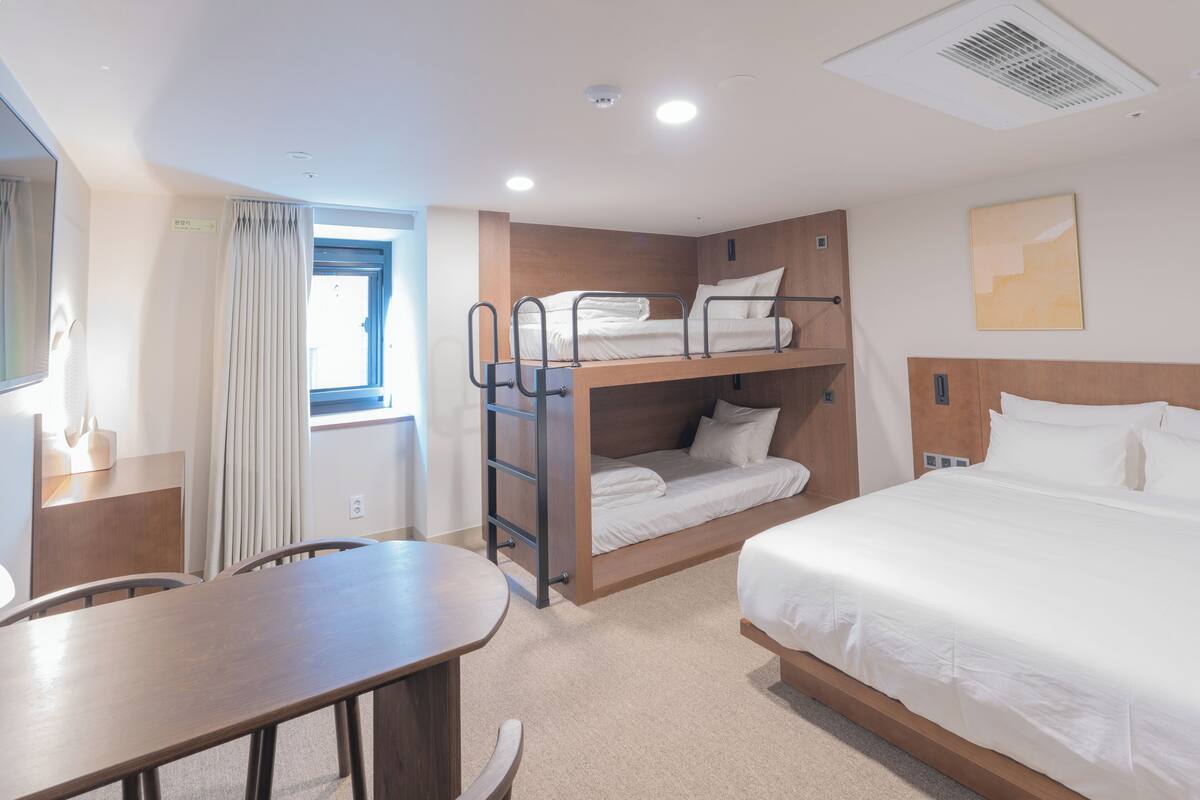 A hotel room with a bunk bed, a single bed, a desk, and a window with curtains.