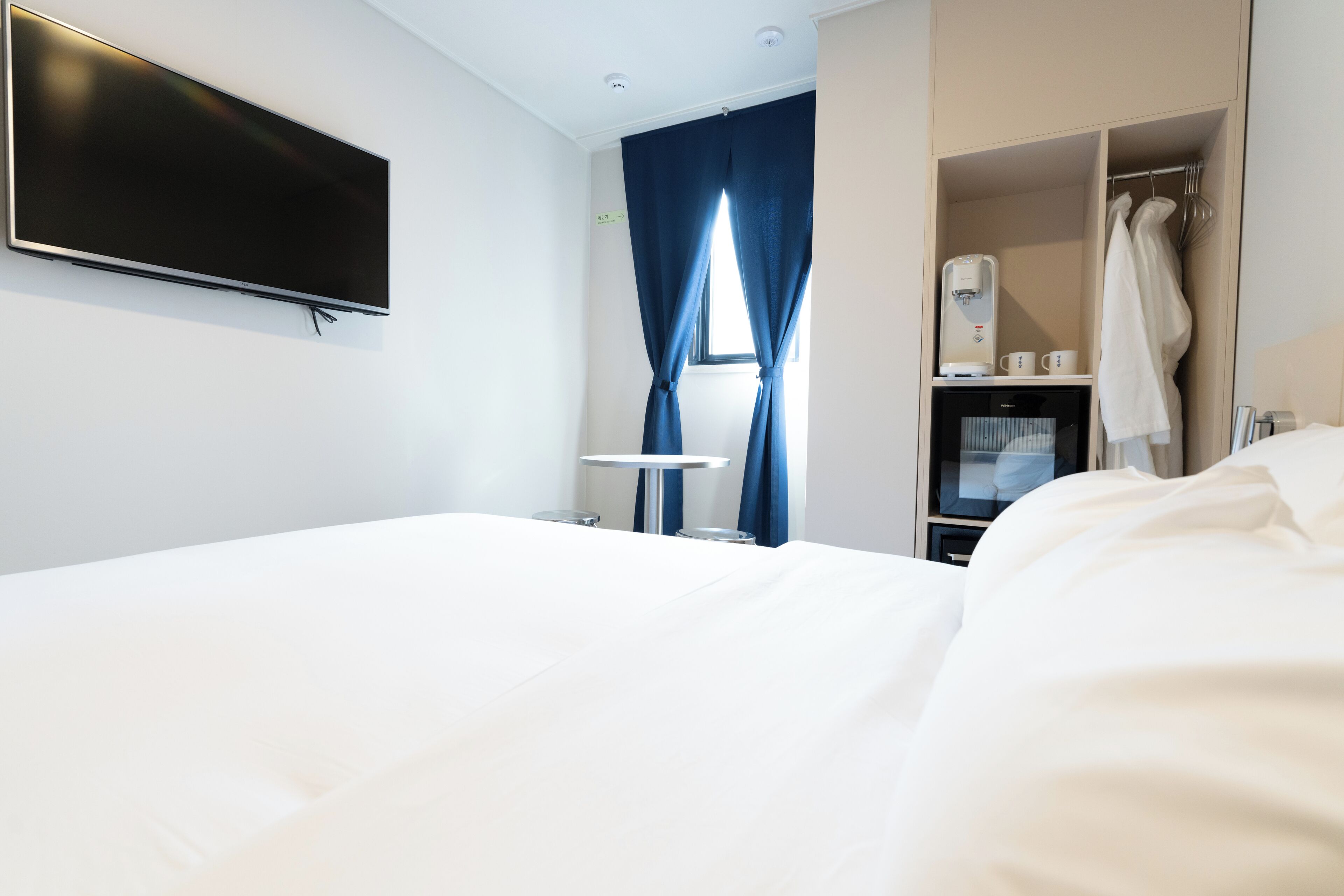 A hotel room with a bed, a flat-screen TV mounted on the wall, a window with blue curtains, a coffee machine, and a wardrobe.