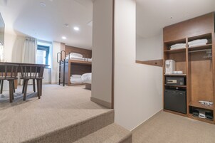 Habitación de hotel con literas, un escritorio y sillas, una kitchenette pequeña y un armario con estantes.
